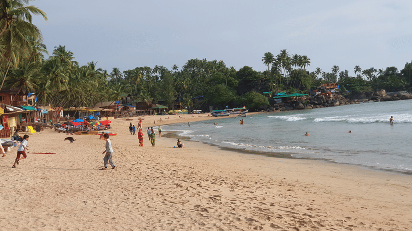 An overview of numerous people strolling on the shores of Majorda Beach during daytime with golden sands, waves overlapping the shoes and trees covering the shoreline.