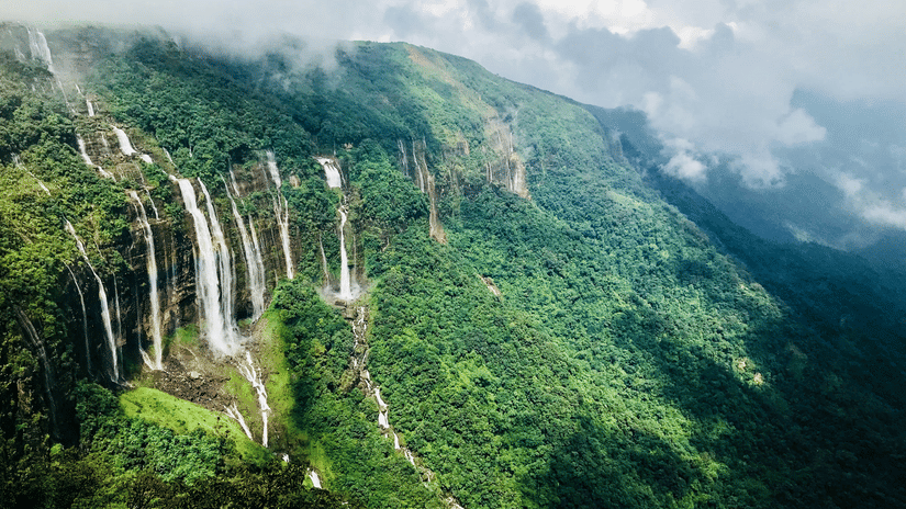 Aerial view of the Seven Sisters Waterfalls cascading down a green cliff face shrouded in mist near Cherrapunji in Meghalaya.