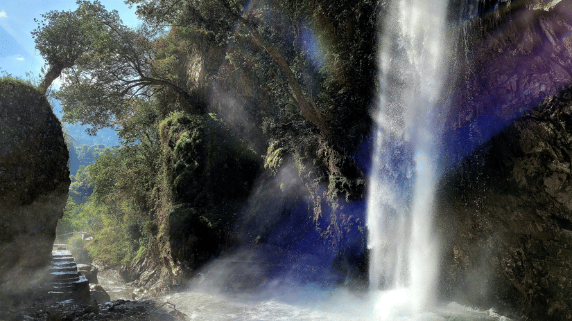 Waterfall plunging into a rocky pool within dense forest, with mist rising and sunlight filtering through trees