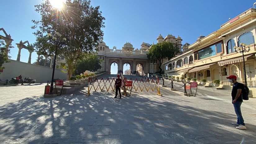 Sunny courtyard with trees and shadows leading to a grand palace entrance, with people walking near the historical site.