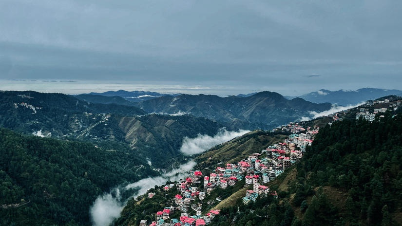 An aerial view of Shimla with many building on the side of a mountain with trees and mountains in the background. Summer is the best season to visit Shimla as the skies are clear and temperate is around 21 degrees.
