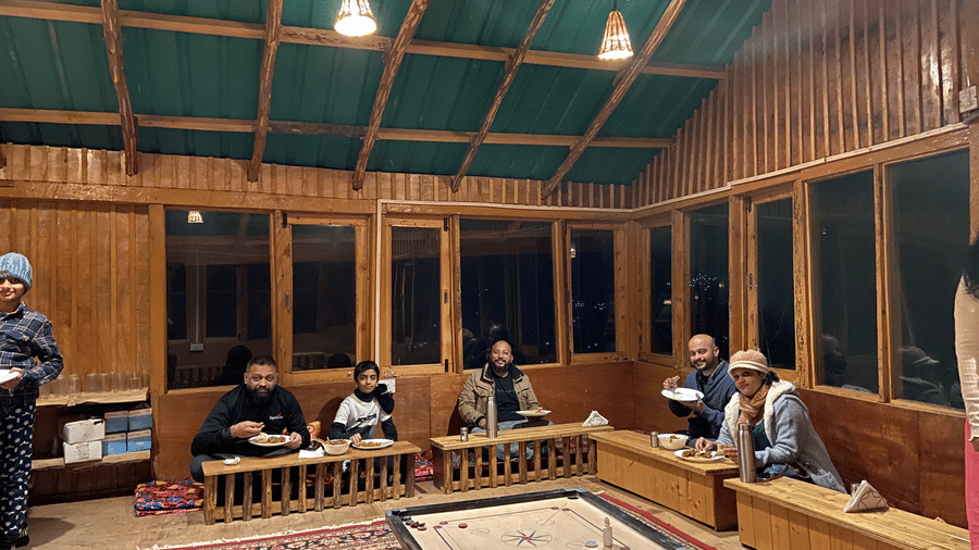 A cosy dining space at Amara Upepo - The Sky Village featuring several people having a meal dressed in winter-wear, along with a carrom board in the middle of the room.