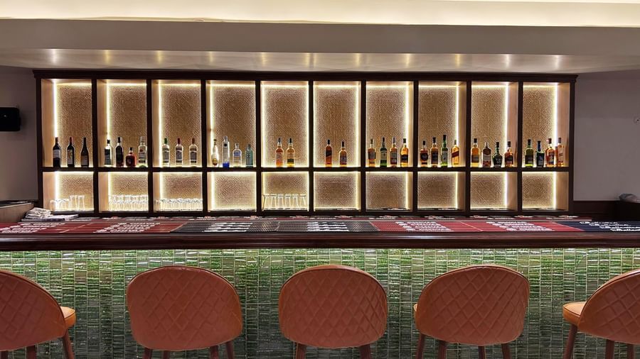  A brightly lit, modern bar counter with an array of spirit bottles displayed in illuminated, wooden cubbies, backed by orange bar stools.