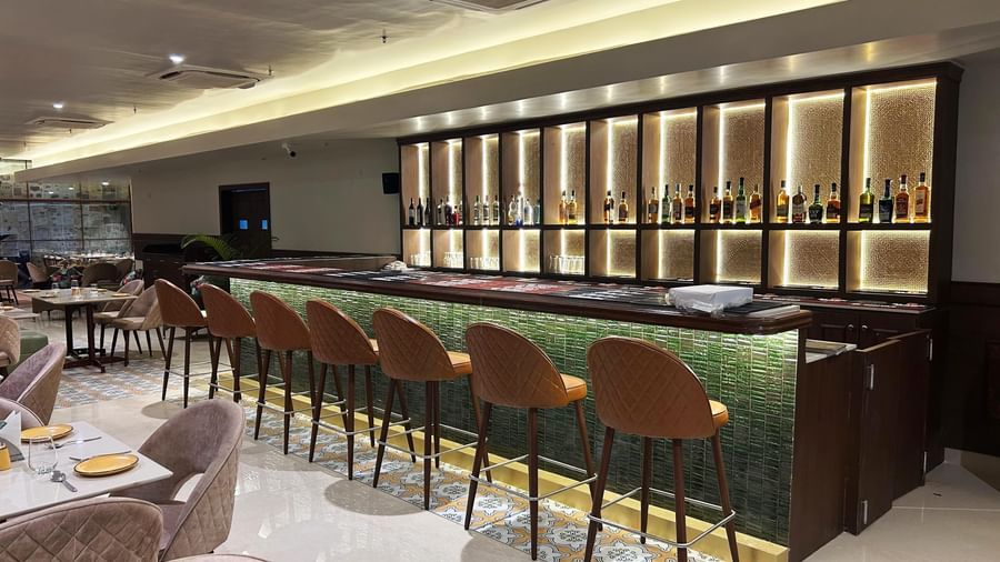 A modern restaurant bar at Amer Greens, featuring an illuminated green bar counter, orange bar stools, and a backdrop of backlit cubbies with spirit bottles.