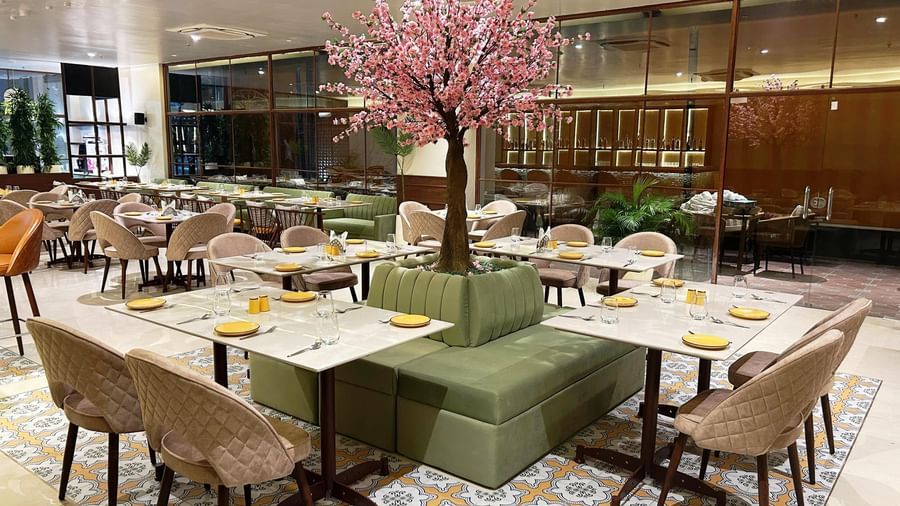 A contemporary restaurant dining area at Amer Greens with a central pink blossom tree planter, surrounded by dining tables and cushioned chairs.