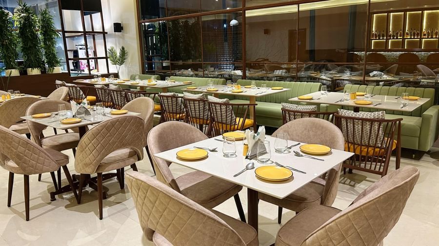  A modern indoor restaurant dining area at Amer Greens featuring square tables with soft chairs in the foreground and a long bench seat along the large window wall in the background.