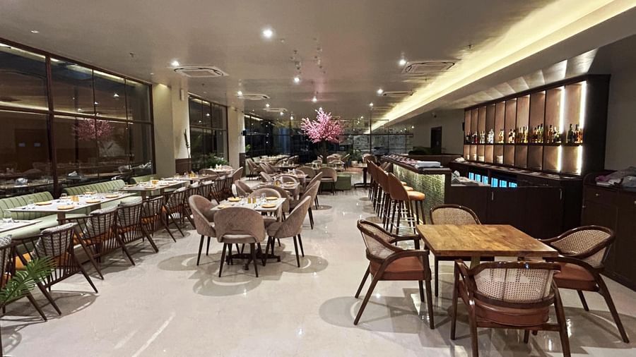 A spacious, modern restaurant interior at Amer Greens featuring multiple dining tables and chairs, with a bar and display area along the back wall.