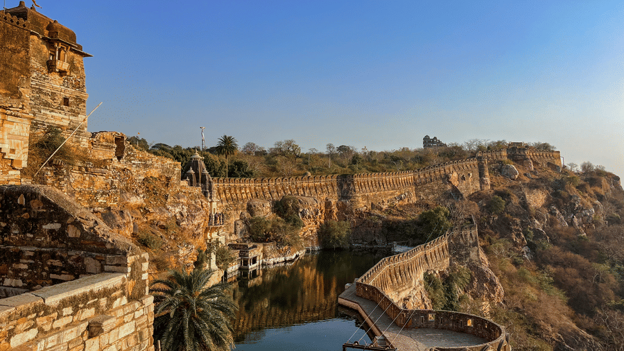 Elevated pathway at Chittorgarh Fort, India’s largest fort, overlooking valley and water body, with stone railings and steep cliffs, offering sweeping views of surrounding rugged landscape