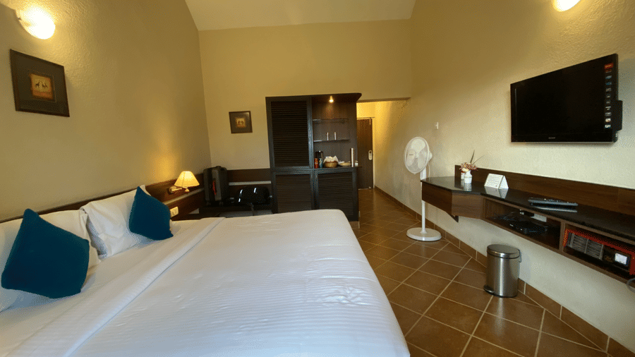 A comfortable and spacious bedroom of Deluxe Room at Green Pastures, Kodai, showcasing a magnificent bed, polished floor tiles, a wall-mounted TV, and a convenient standing fan near the foot of the bed.