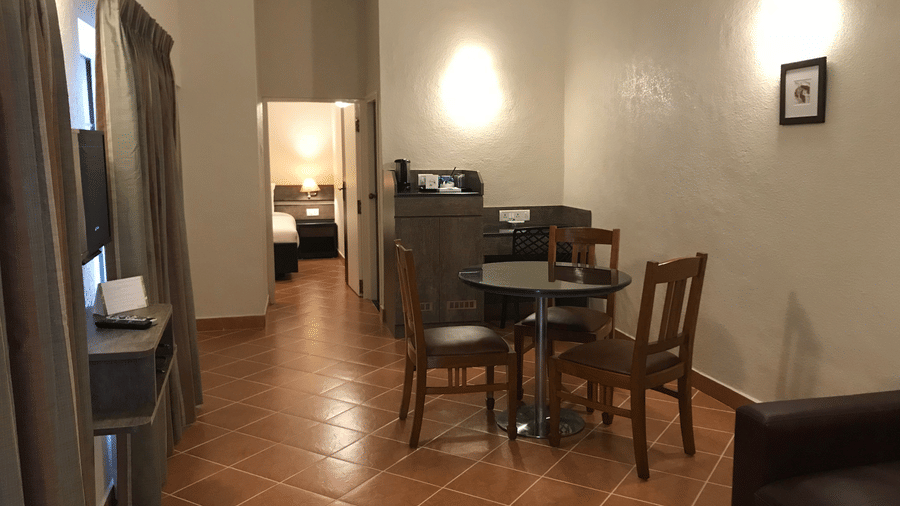 The Inviting Luxury Suite living space at Green Pastures, Kodai, featuring polished tile flooring, a sophisticated dining set for four, a comfortable armchair, and an extended view down a pleasant hallway toward the bedroom.