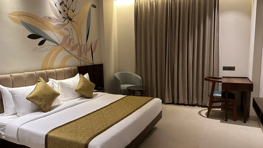 A well-appointed bedroom with a king size bed featuring a padded headboard, neatly arranged pillows, a bed runner, wall art above the bed, a lounge chair beside a side table, full-length curtains, and a work desk placed near the window at Hotel Bawa, Udaip
