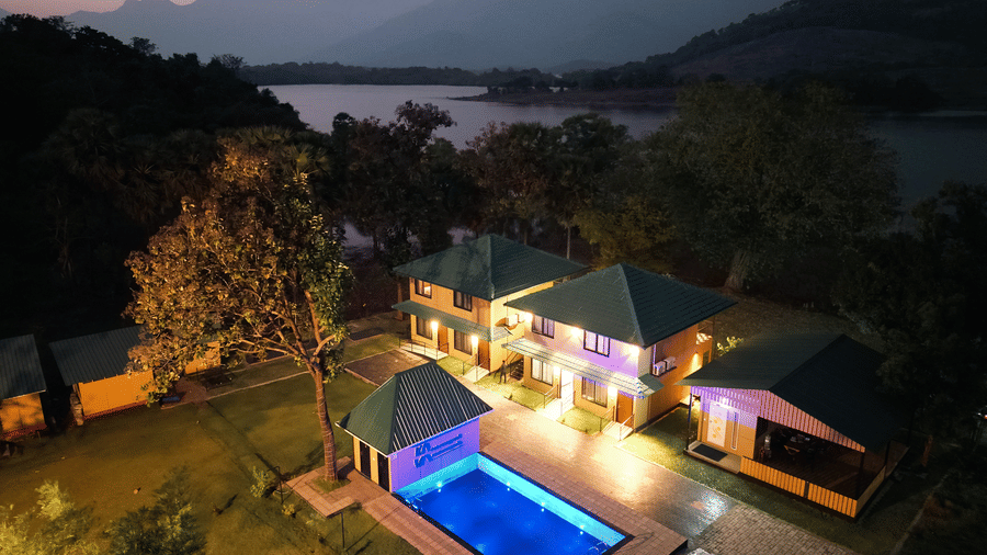 High-angle night view of the resort showing the glowing blue swimming pool, lit pathways, and the dark silhouette of the lake and mountains - Ibex Resorts, Malampuzha (Kava Eco Camp and Caravan Park)