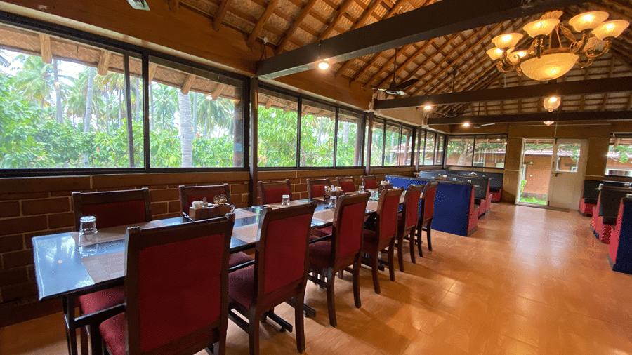 Elegant dining area at Ibex River Resort, Pollachi with neatly arranged wooden tables and chairs, large windows offering views of the surrounding greenery, and warm ambient lighting.