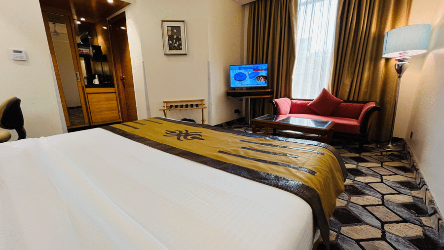 A bedroom at JRD Exotica - Business Hotel featuring a neatly-made bed, a patterned carpet, a sofa, and full-length curtains.