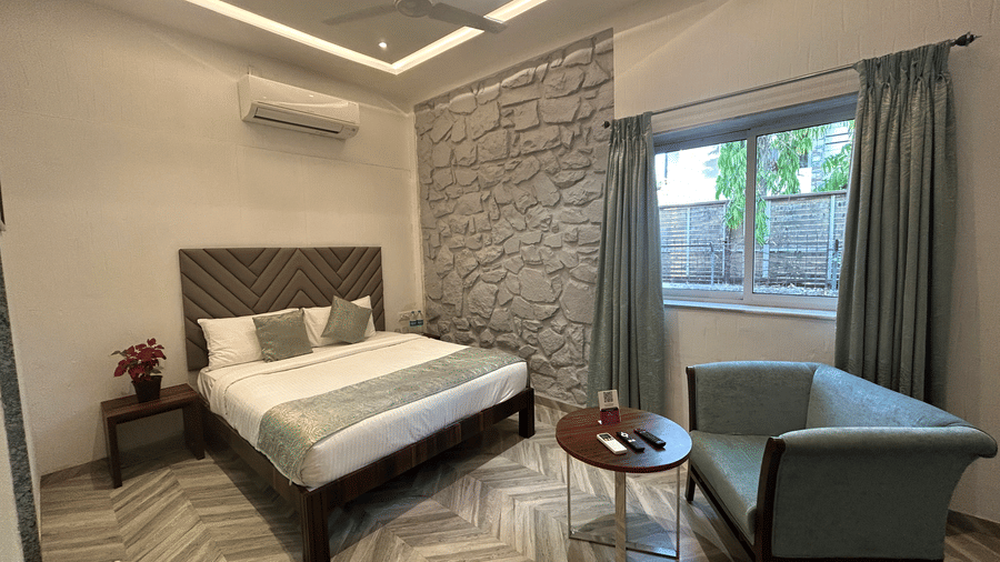 A bedroom featuring a plush king size bed, elegant stone-textured walls, and a cosy seating area, illuminated by natural light streaming through large windows – Maxxvalue Klub Klass Villa, Lonavala.