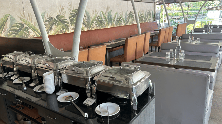 A well-arranged buffet dining setup with stainless steel chafing dishes, plates, and cutlery, set in a semi-outdoor setting– Maxxvalue Klub Klass Villa, Lonavala.