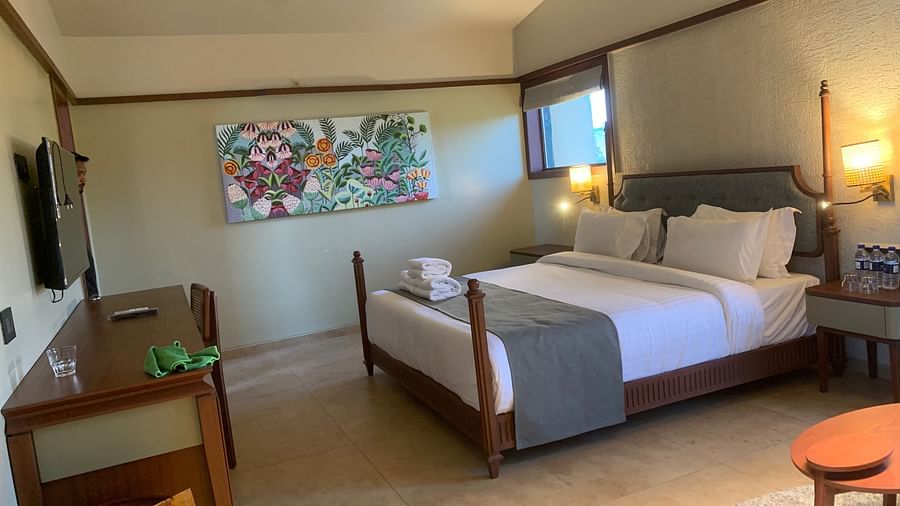 Side view of a double bed with grey bedsheet, a small round table and a beautiful painting on the wall at Pride Resort near Ambaji Temple.