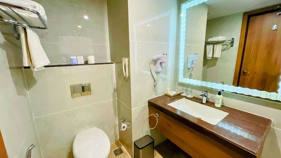Modern hotel bathroom featuring a wall-mounted toilet, towel rack, large mirror with built-in lighting, spacious countertop with sink, and neatly arranged toiletries - Sobit Sarovar Portico, Goa.