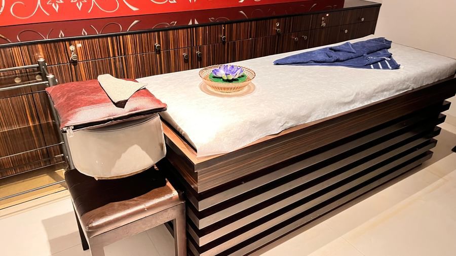 A neatly arranged spa therapy room with a cushioned massage table, folded towels, floral decor, and warm red patterned wall design at Springs Hotel & Spa, Bangalore