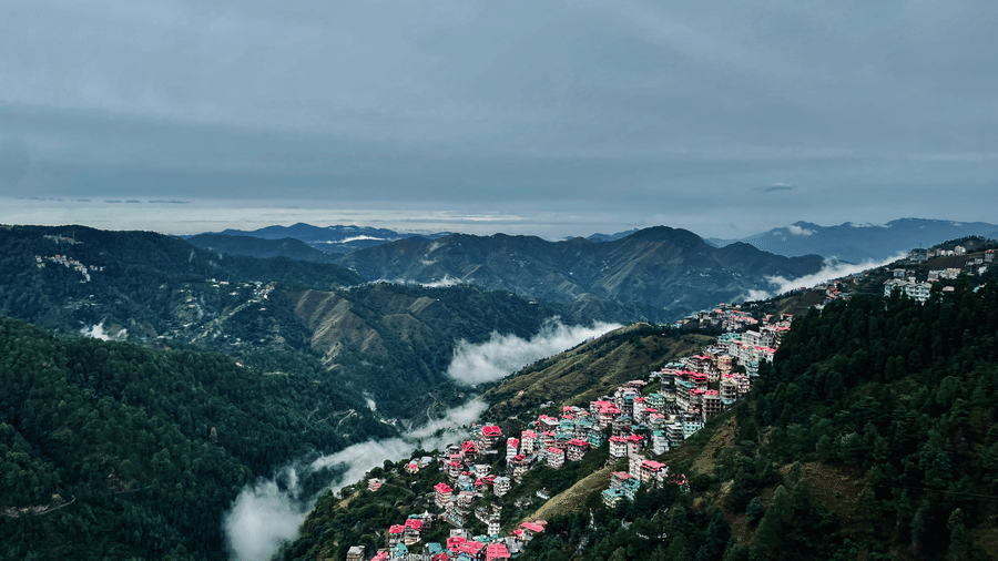 An aerial view of Shimla with many building on the side of a mountain with trees and mountains in the background. Summer is the best season to visit Shimla as the skies are clear and temperate is around 21 degrees.