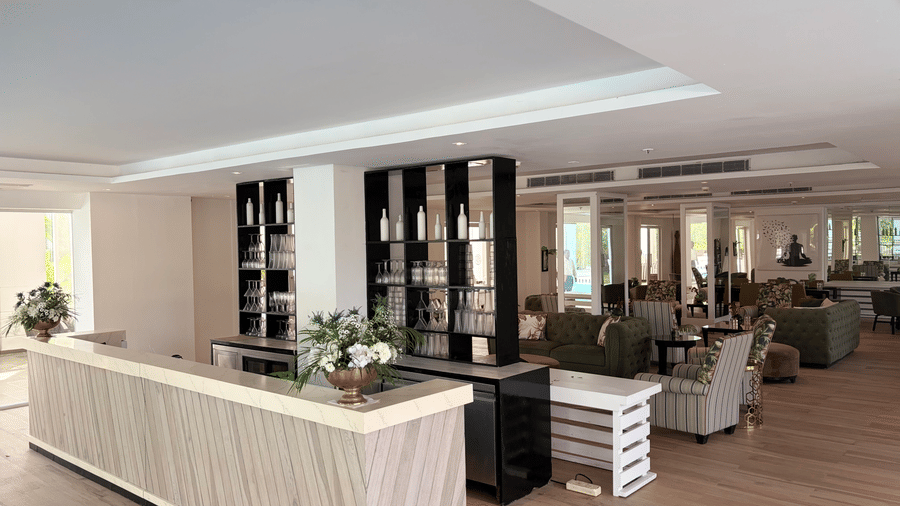 Indoor bar and lounge at The Mansion House resort with light wood counter, black shelving with glassware, green and grey sofas, and recessed ceiling lights.