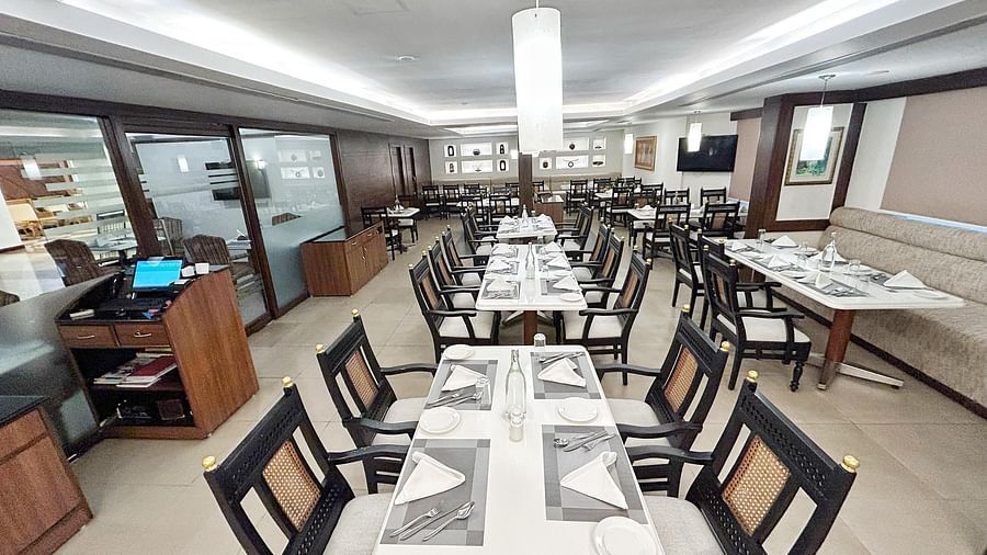 A well-lit, empty restaurant with neatly arranged tables, dark chairs with light cushions, and a long central light fixture at The Residency Karur.
