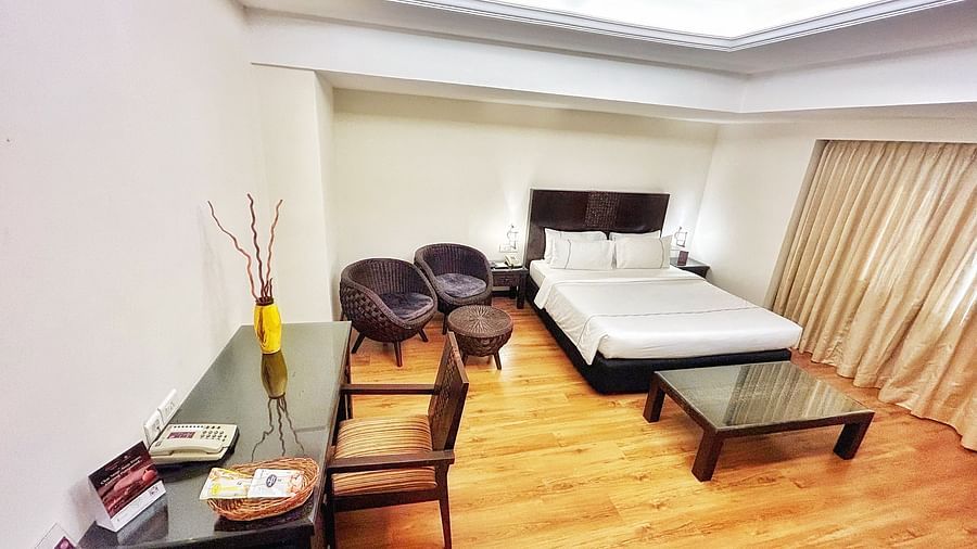 Residency Suite at The Residency Karur shows a double bed, a glass coffee table, two round chairs, and a desk with a wooden chair on a wooden floor.