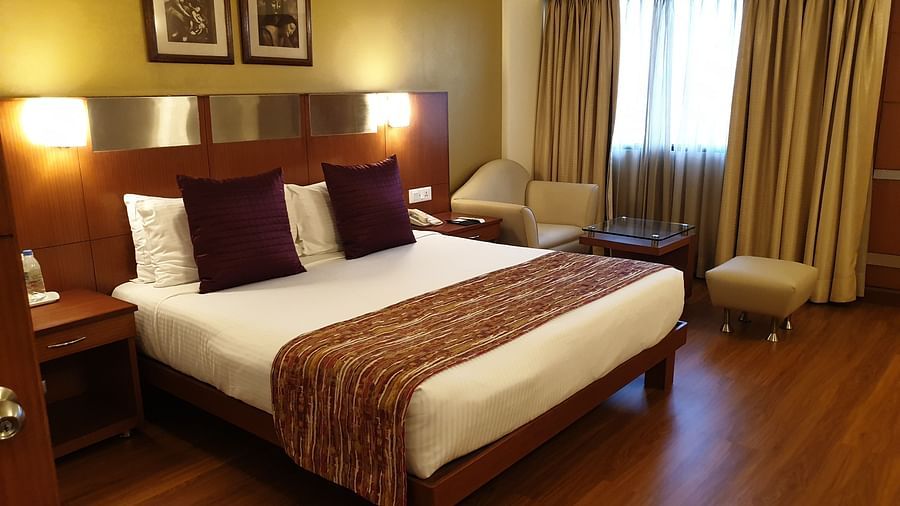 An image of a Deluxe Room with double size bed, bedside table and lam on either side and a huge window covered with curtains - The Suryaa New Delhi