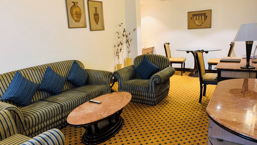 An image of a seating area with sofa sets, centre table, flat TV screen, and a cabinet in a Deluxe Suite - The Suryaa New Delhi