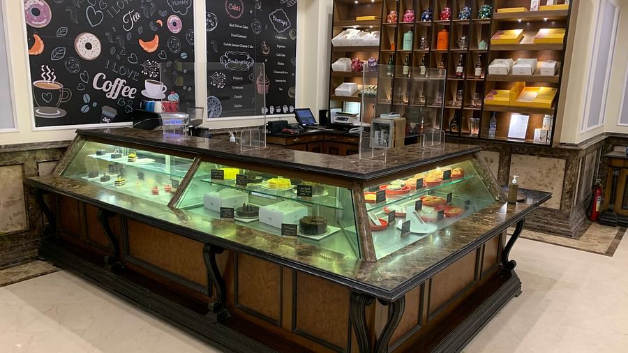 An image of a cafe where at the front desk cakes are kept which can be seen through the glass and on the wall a cabinet is attached where  liquor bottles and pots are kept - The Suryaa New Delhi