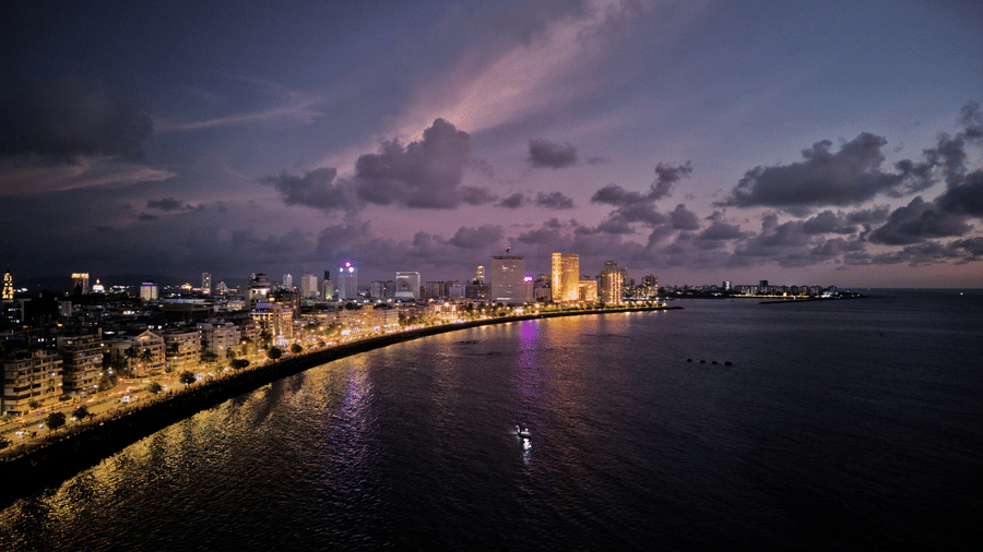 Evening view of a coastal city promenade with illuminated buildings and reflections across the waterfront