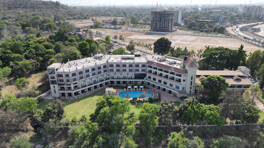 An aerial view of VITS Kamats Resort, Silvassa, a resort in Silvassa for weddings, surrounded by trees with a swimming pool, a lawn area in front of it and a highway, open land and an under-construction building in the background.