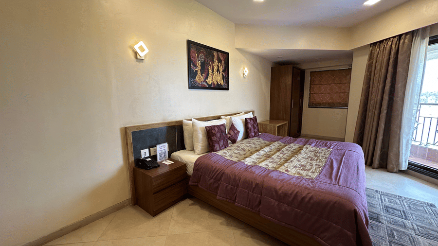 A side view of the Riviera Suites with a double bed, side tables having storage, and ceiling-to-floor windows opening to the balcony at VITS Kamats Resort, Silvassa.