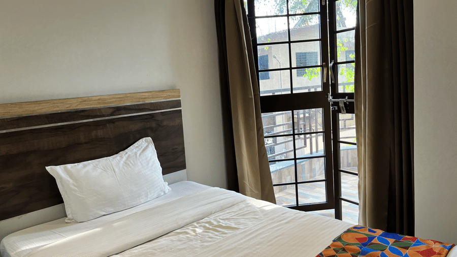 A close-up of a single bed near a glass & wooden door to the balcony, with green curtains next to the door inside Classic Single Room - the best hotel room in Kharadi at VITS Select Kharadi, Pune.