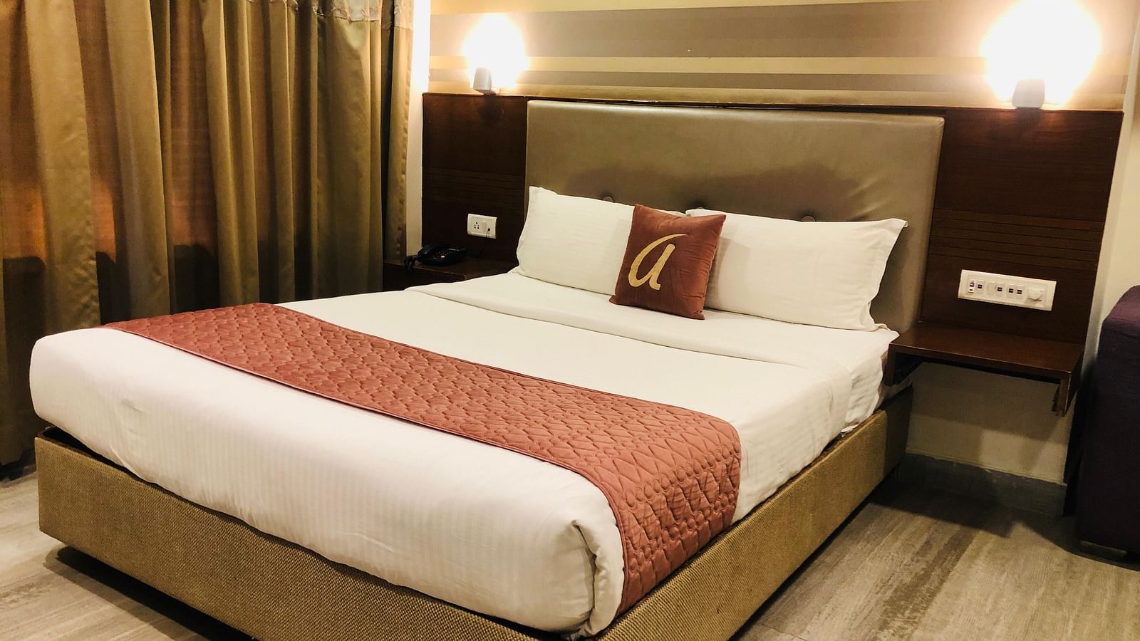 Hotel room double bed with white linens, maroon runner, and decorative pillows, illuminated by soft headboard lighting | Astra Hotels and Suites Kadubeesanahalli