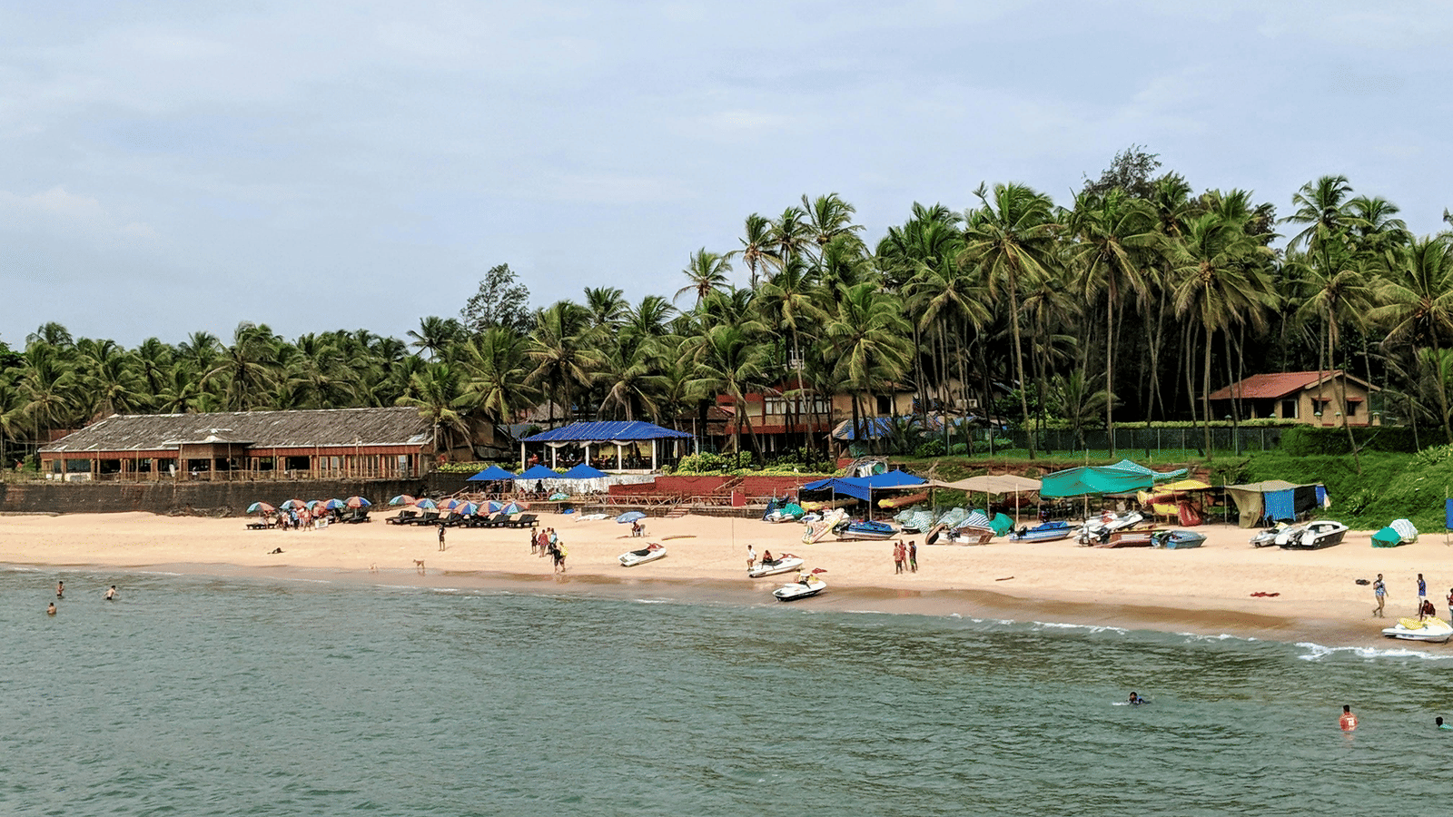 A far-away view of a beach with a many boats, coconut trees, people swimming in the beach, and a beach shack in the distance. Visiting a beach shack is one of the best things to do in Goa in monsoons.