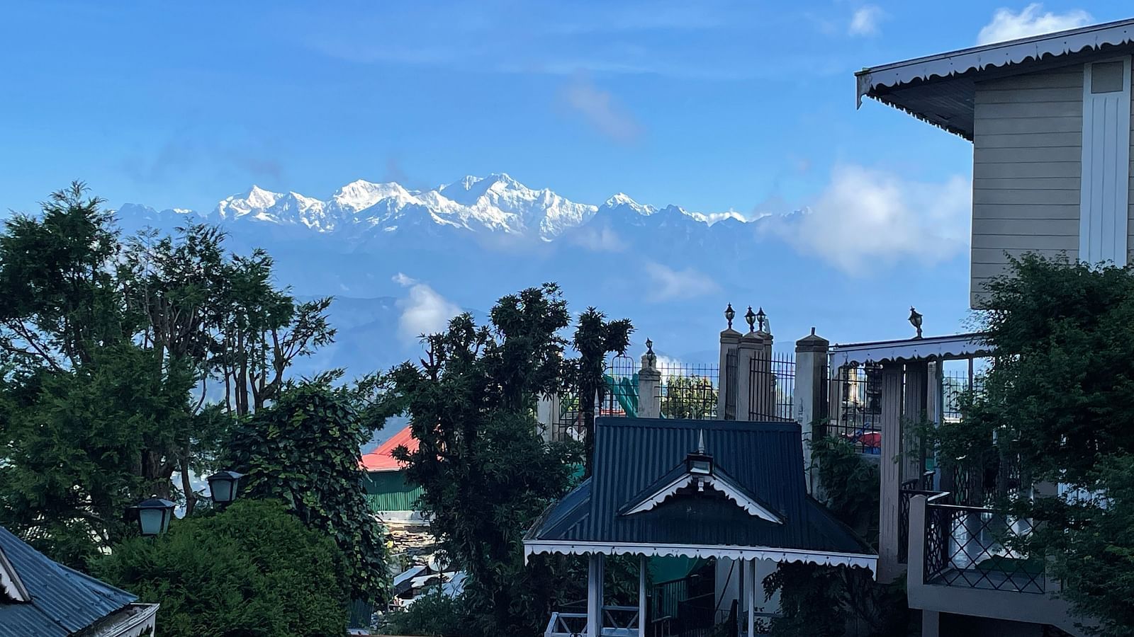 View of snow-capped Himalayan peaks in the distance from the peaceful garden area at Cedar Inn, Darjeeling, framed by rooftops and lush greenery.
