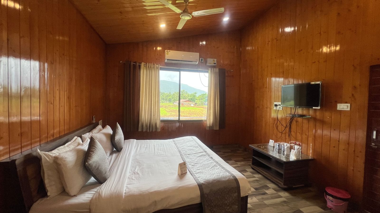 Side view of Daksh Elite Cottages showcasing it's double bed facing a wall-mounted TV, and a window beside offering views of the surroundings.