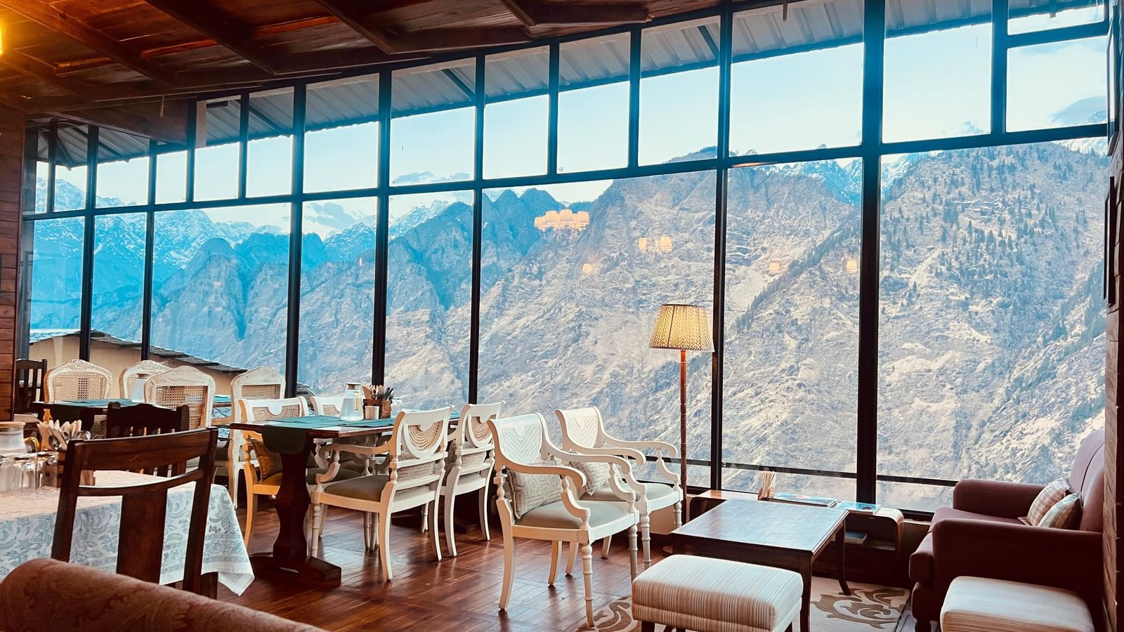 Dining area at Faraway Cottages, Auli, with large windows revealing a mountain range under a clear blue sky, for the perfect Stay Near Badrinath Temple.