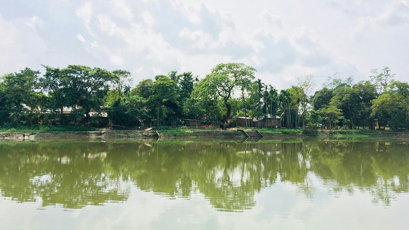 Serene lake with trees and greenery reflecting on the calm water under a cloudy sky.