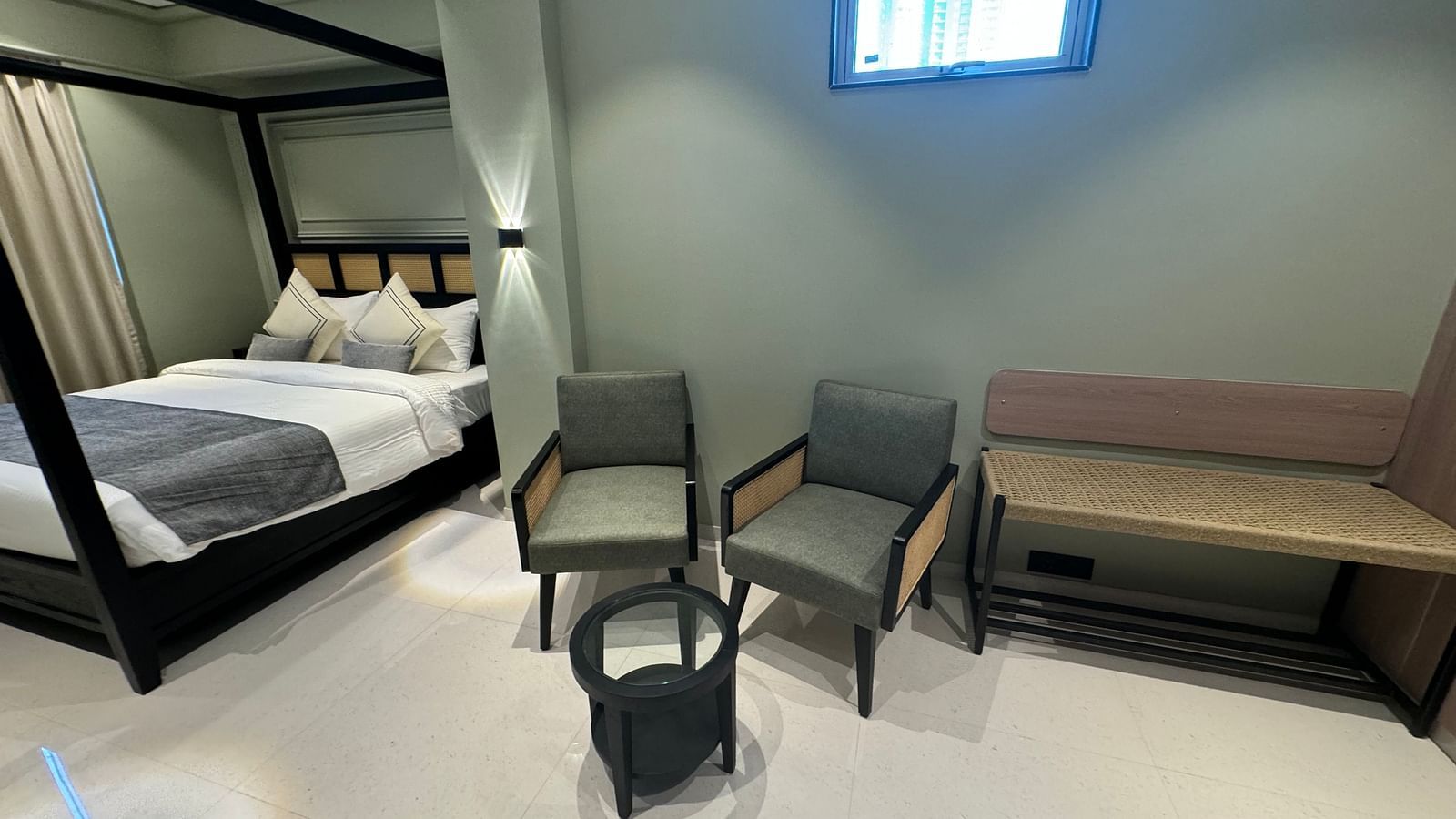 A modern hotel room featuring a black four-poster bed with white bedding and decorative pillows, two gray armchairs, a black round coffee table, and a wooden bench against a wall with a high window.