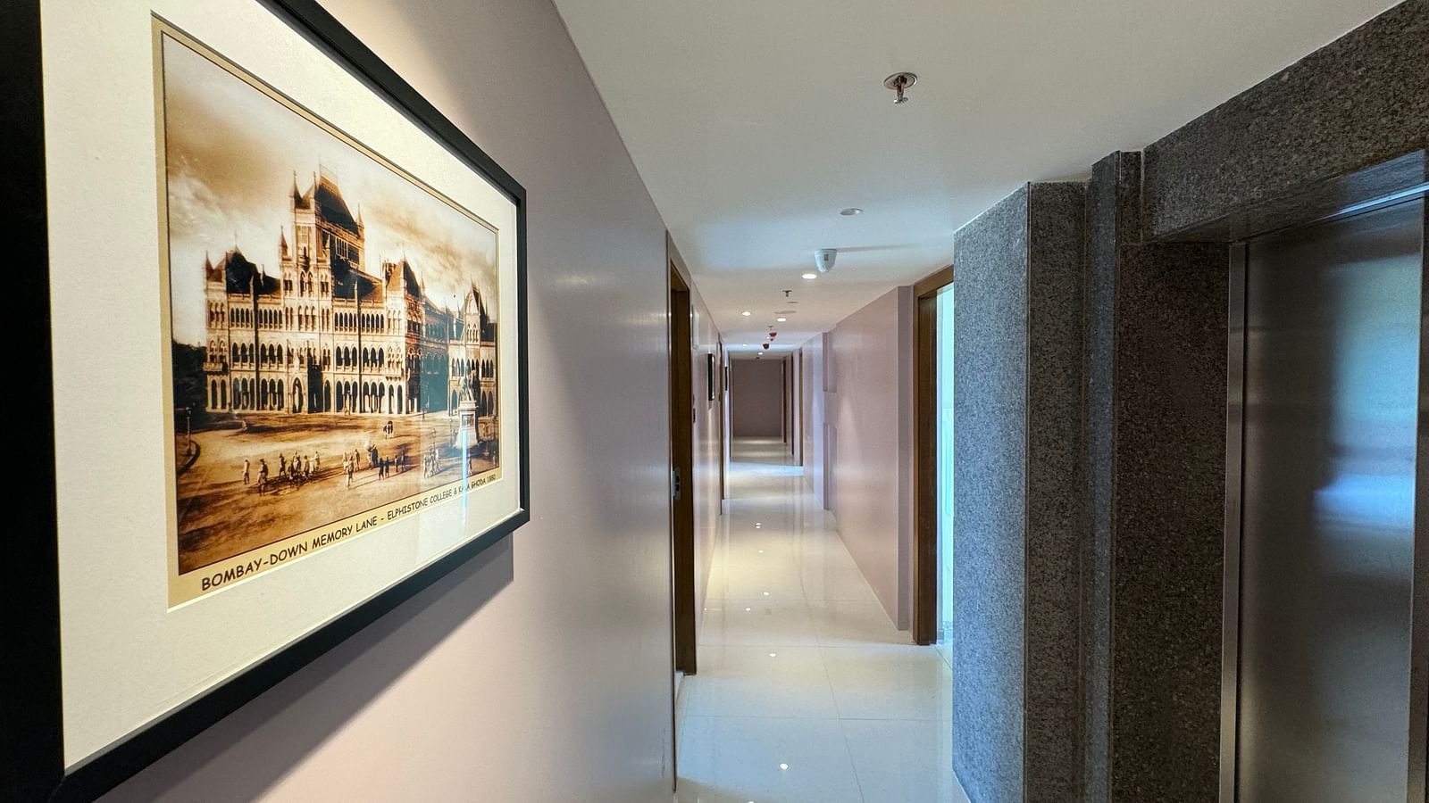 A corridor in a hotel with a framed picture on the wall showcasing historical architecture, glossy tiled floors, and ambient lighting.
