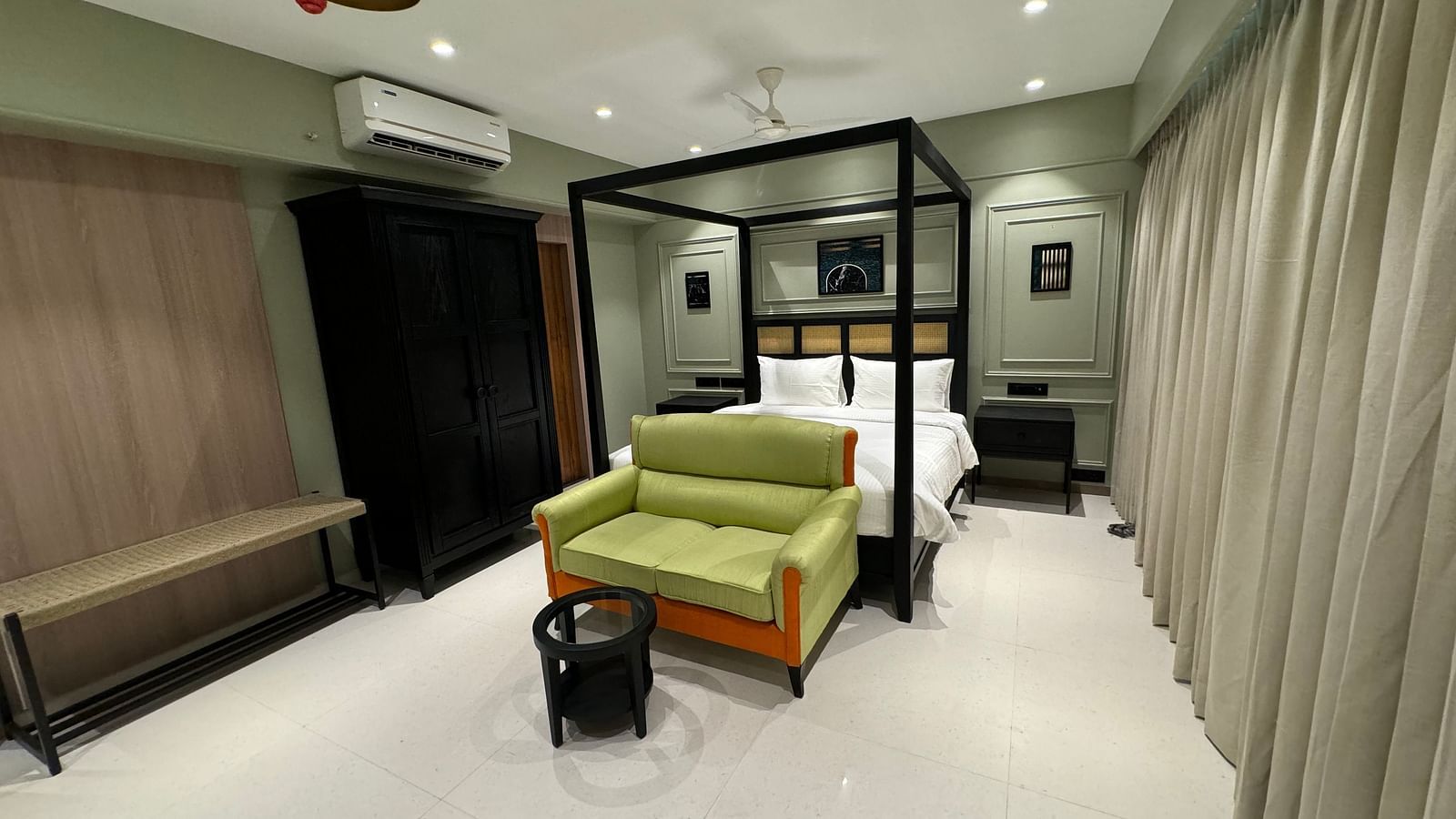 A hotel room with a black four-poster bed and white bedding, a green sofa, a black coffee table, a woven bench, and a black wardrobe, illuminated by ceiling lights and lamps.