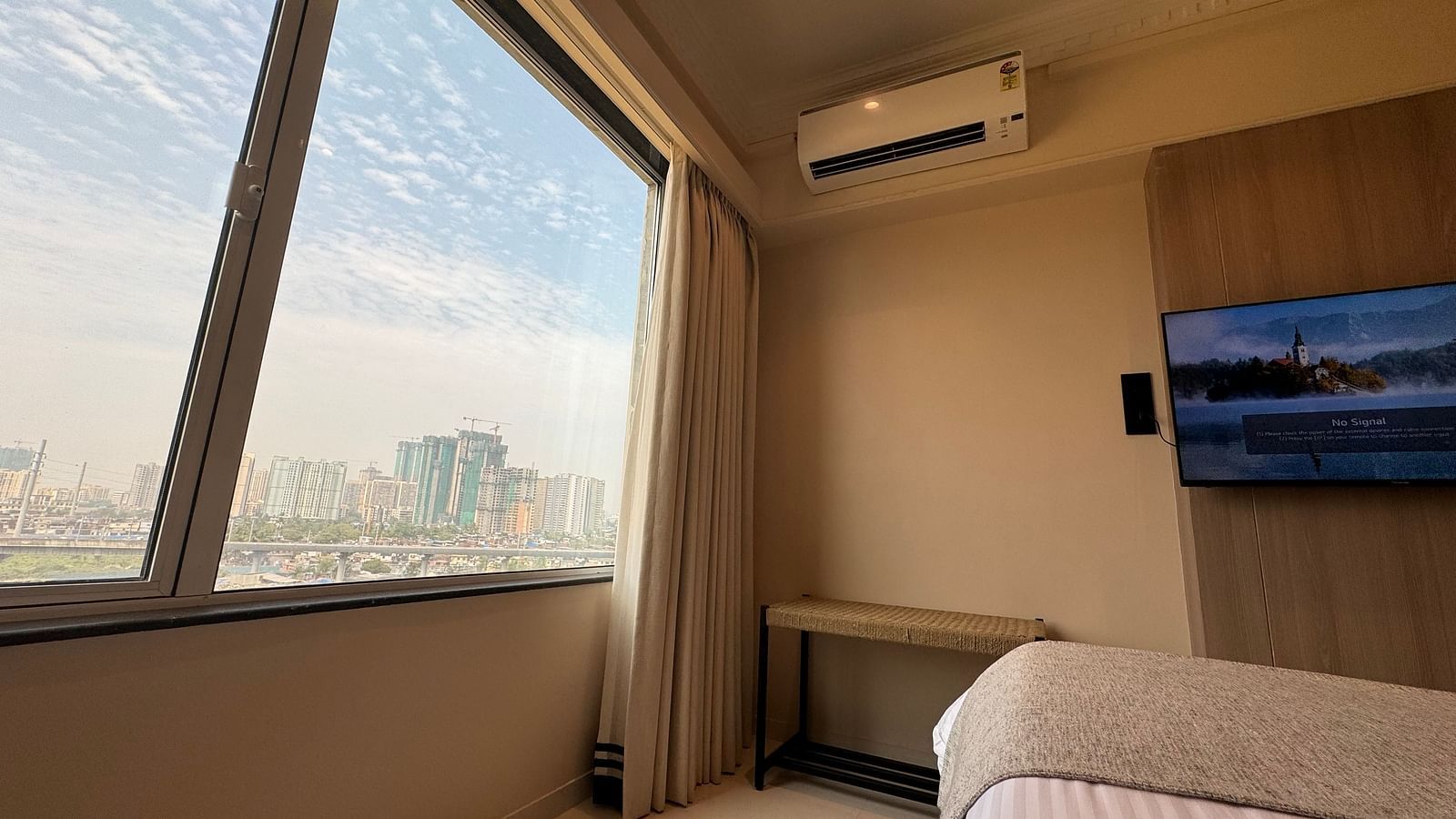A city-view room with a bed and TV, simple decor, and a large window with a blue sky outside.
