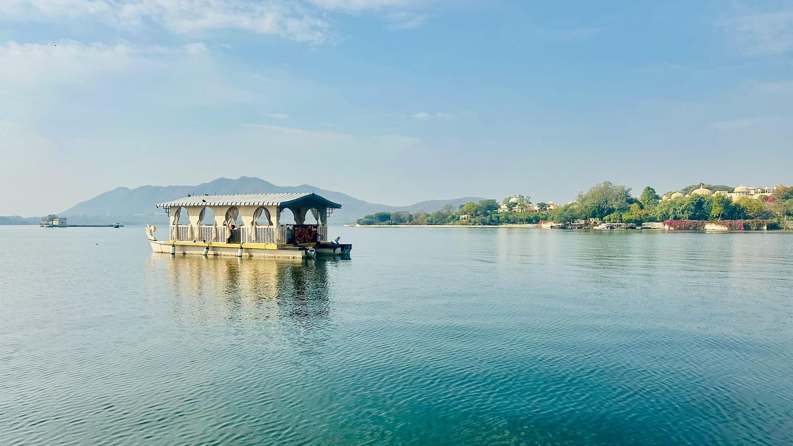 A traditional houseboat floats peacefully on the still, blue waters of a lake, with distant green hills under a clear sky.