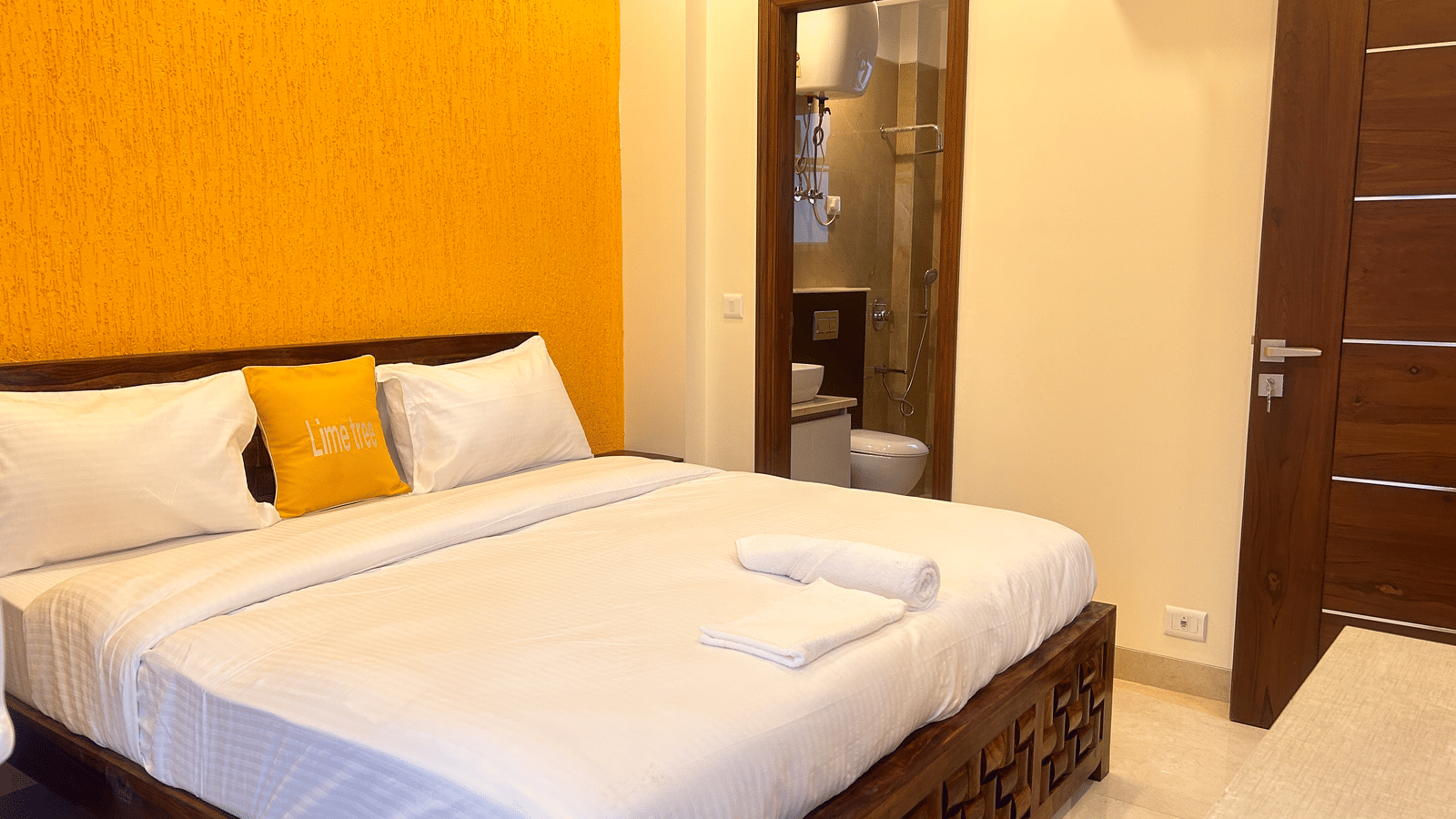 A bedroom with a yellow accent wall, white bedding, wooden furnishings, and an attached bathroom visible through the door - Lime Tree Hotels & Banquet, Vrindavan