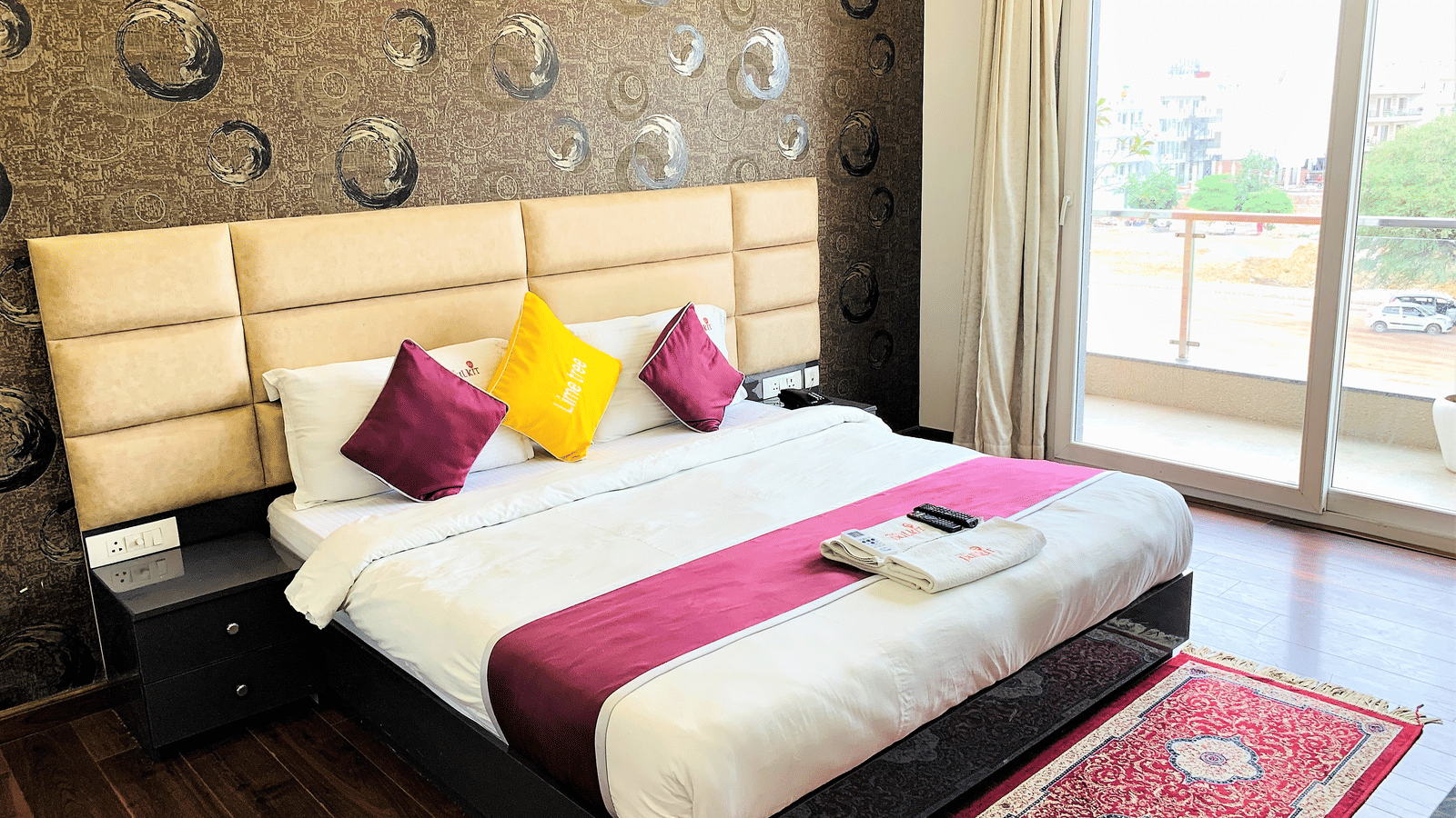 An image of a bedroom with a double size bed, a bedside table, and a glass door which leads to the balcony - Lime Tree Hotel. Pulkit