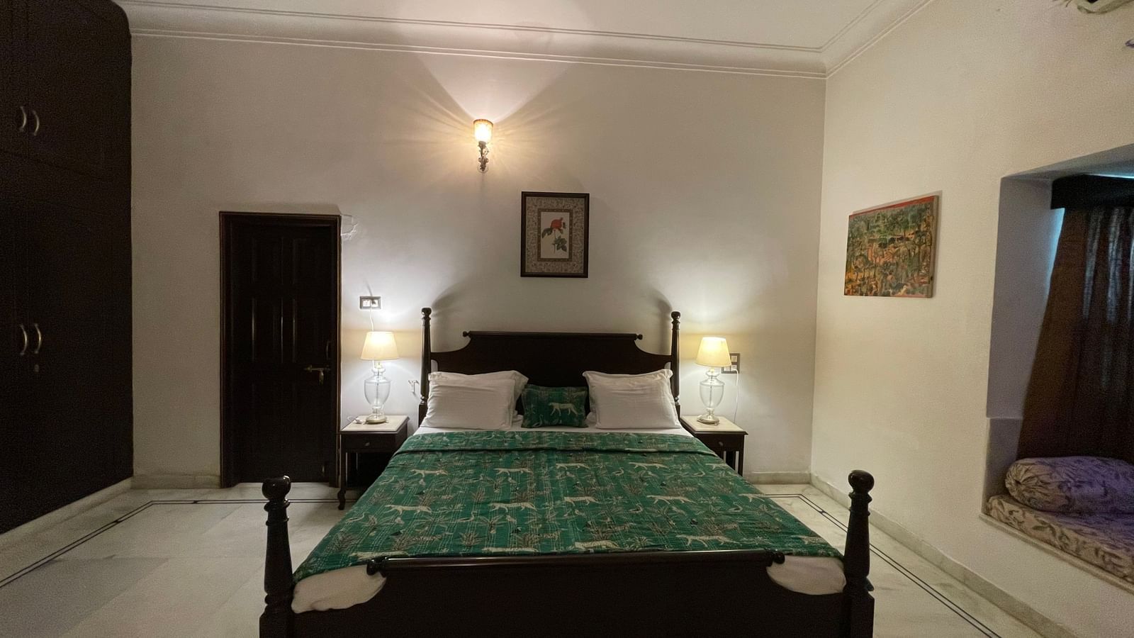 An eye-level view of the heritage room at Mahendra Niwas, showcasing the bed and a dark wooden armoire, with traditional paintings on the walls and warm lighting.