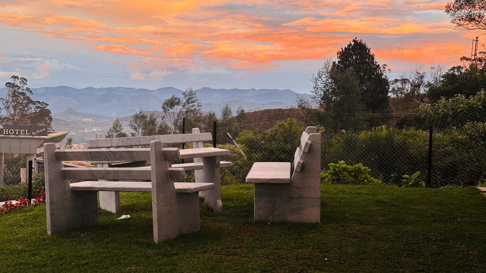 An outdoor concrete table and benches on a grassy hillside, overlooking a valley at sunset, at MYST Resorts, Doddabetta, Ooty.