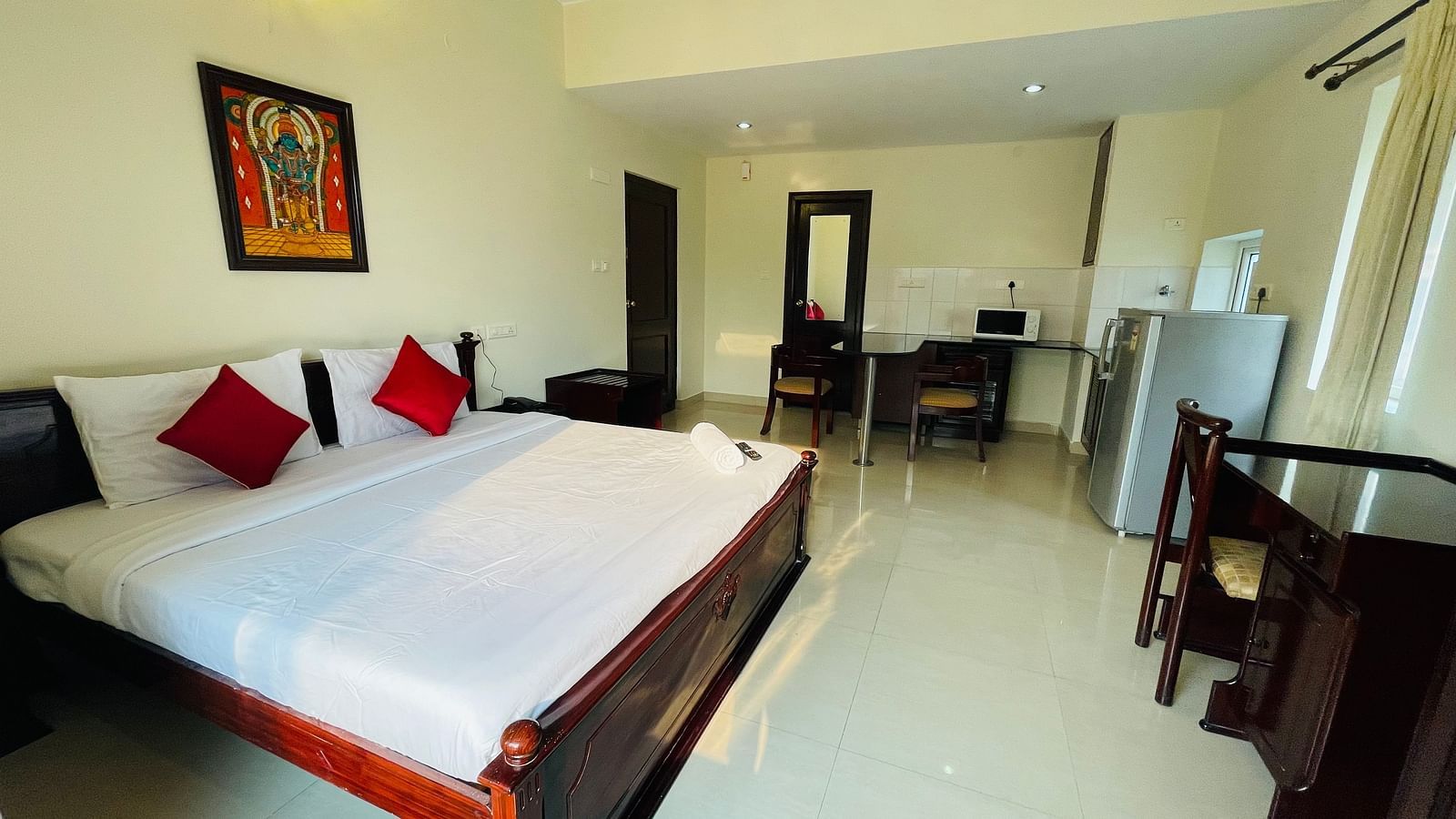 A room with a double bed featuring white linens and red cushions, a separate seating area, and a glimpse into a kitchenette space at Sanctum Suites Indiranagar, Bangalore.